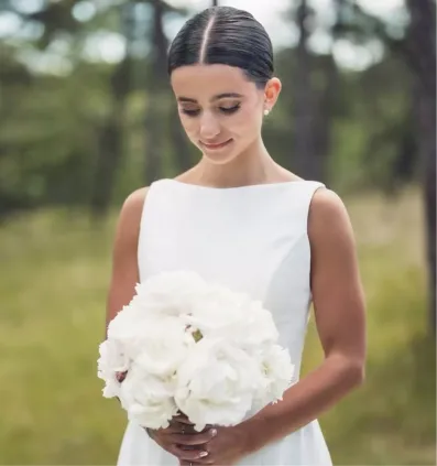 Woman in a white sleeveless dress holding a bouquet of white flowers, outdoors with blurred trees.