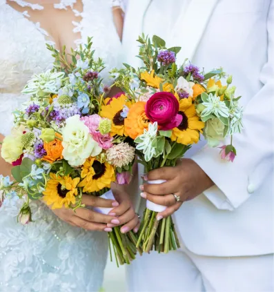 Two people in wedding attire holding colorful bouquets with sunflowers and ranunculus flowers.