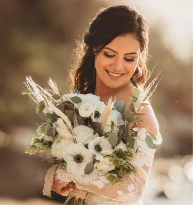 Smiling bride holding a bouquet of white flowers and greenery in soft natural light.