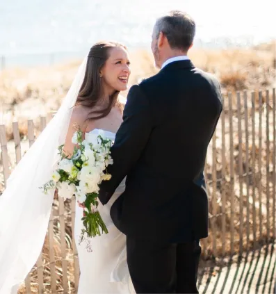 Bride in white dress holding bouquet smiles at groom in black suit by a wooden fence outdoors.
