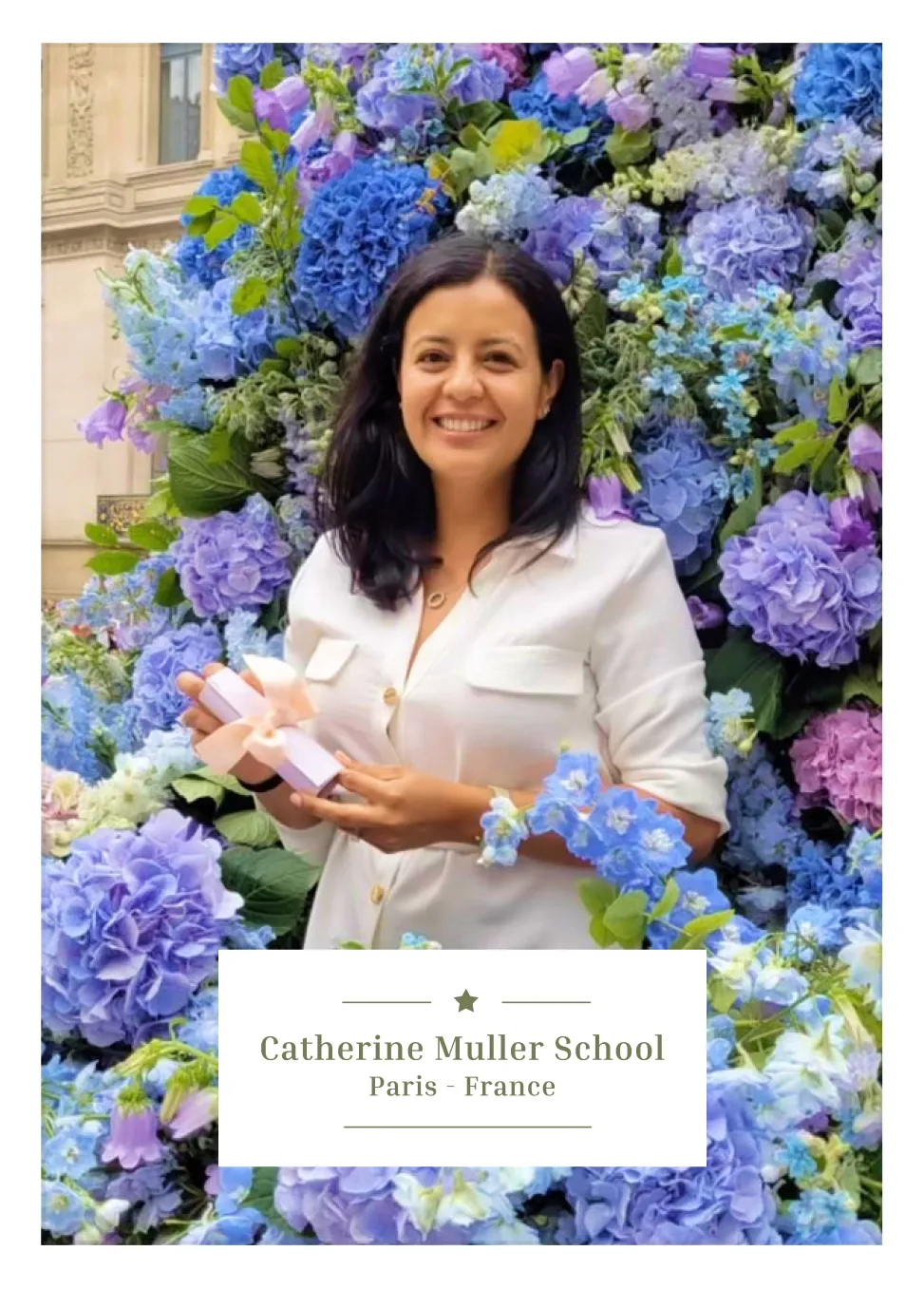 Smiling woman in white blouse holding a small gift box, standing in front of blue and purple flowers.